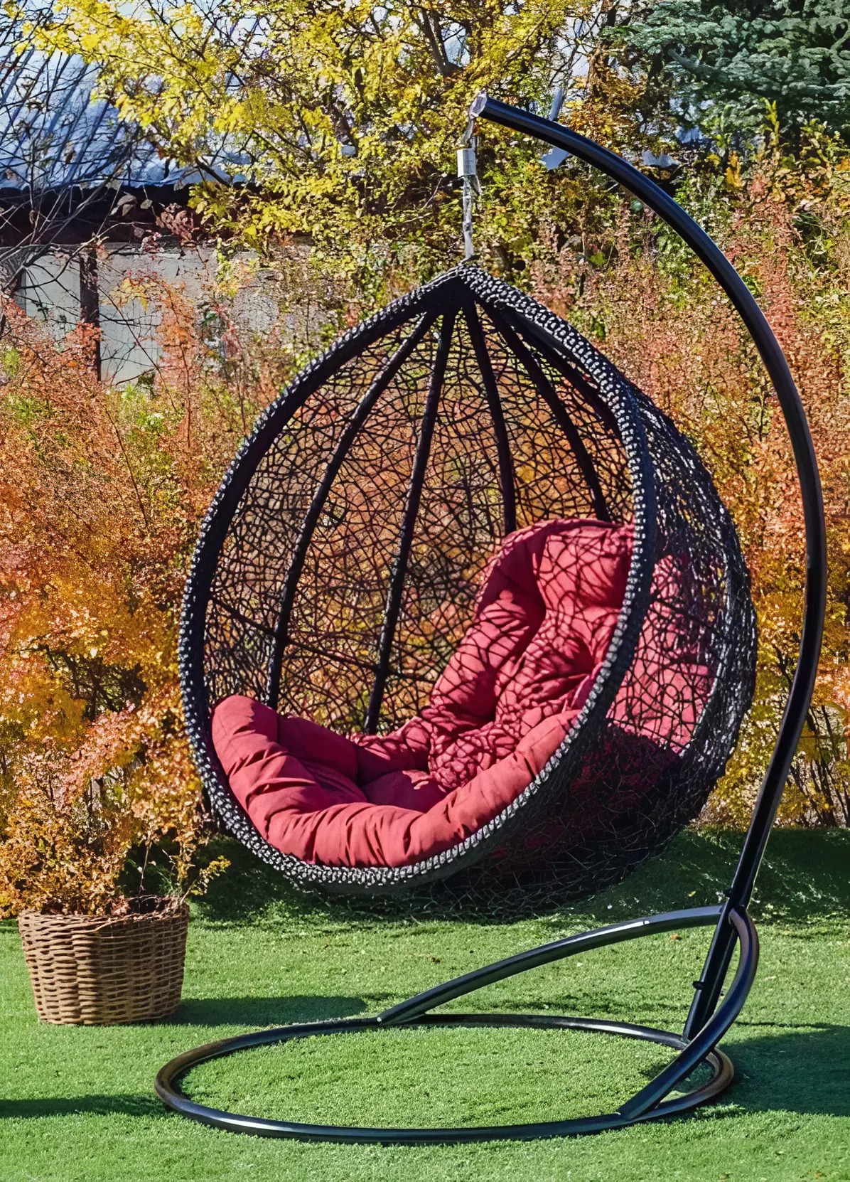Luna hanging egg chair in black rattan with a red cushion, hanging in a garden with autumn foliage behind it.