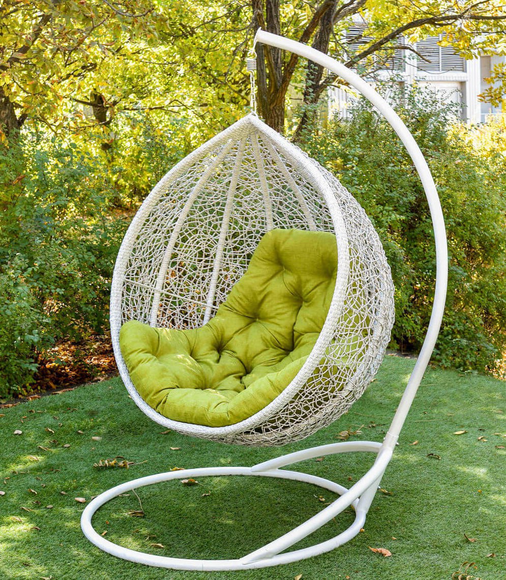 Luna hanging egg chair in white synthetic rattan with an olive green cushion, hanging from its white metal stand on a lawn.