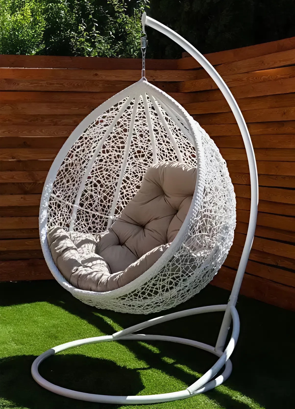 Luna hanging egg chair in white rattan with a taupe cushion, set up on artificial turf beside a timber fence.